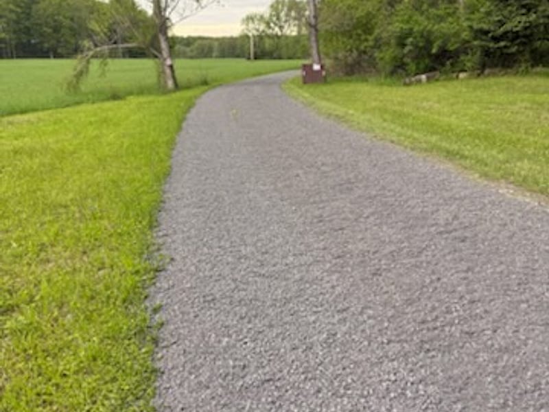 A long crushed-stone driveway running between a hayfield and a treeline
